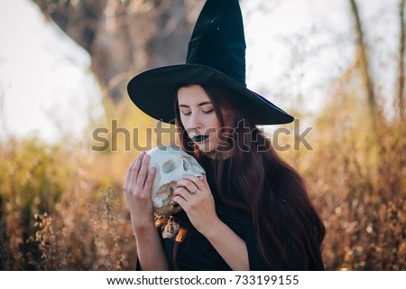 A young witch with pale skin and black lips in a black hat, a dress and a raincoat. Holds the skull of a dead person. Against the background of a tree of gallows. halloween, magic, fantasy, necromancy