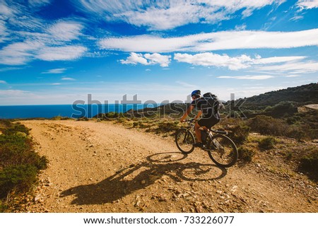 a young guy riding a mountain bike on a bicycle route in Spain on a dirt road against the background of the Mediterranean Sea. Dressed in a helmet, a dark one and a black backpack