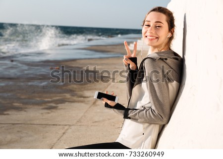 Close-up photo of happy sport woman showing peace sign, looking at camera while listening to music