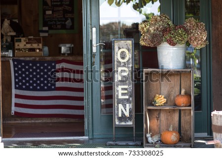 A small business is open for business with a welcoming open sign, pumpkins and dried flowers. Inside the store hangs an American flag. 