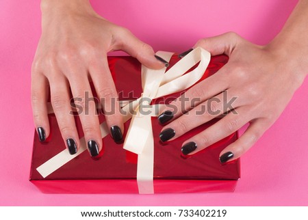 Female hand with black nails manicure on red gift box with white bow on pink background, close up