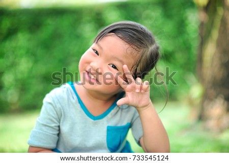 A little cute asian girl show hand in shape of fighting near eye and smile on the green grass background. (Selective focus)