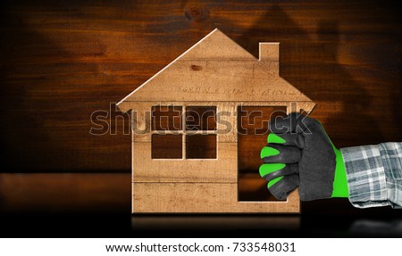 Construction industry concept - Hand with work glove holding a wooden model house on a table with reflections