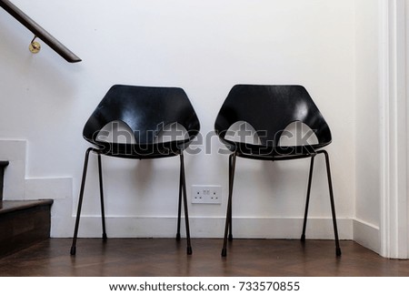 Two black retro chairs standing against a white wall, with dark wooden floor. Photographed October 2017.
