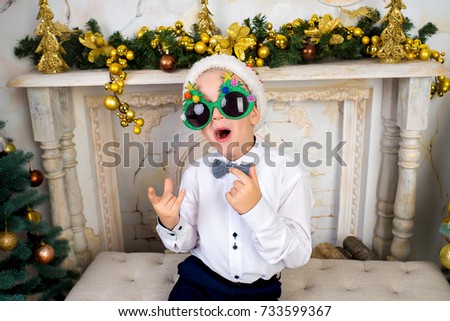 Creative funny boy seating on white chair at Christmas time. With colorful lights from Christmas tree, fireplace on background, selective focus. Horizontal banner, conceptual idea for New Year party