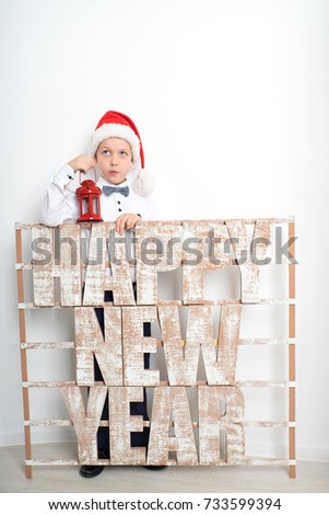 Smiling cute boy standing near white studio bricks wall at Christmas time. Santa Claus hat, red candlestick lights for Christmas. Large signboard with big three dimensional letters Happy new year