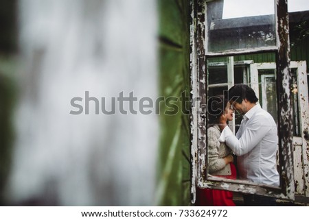 Man kisses pregnant woman in grey sweater standing before an old window