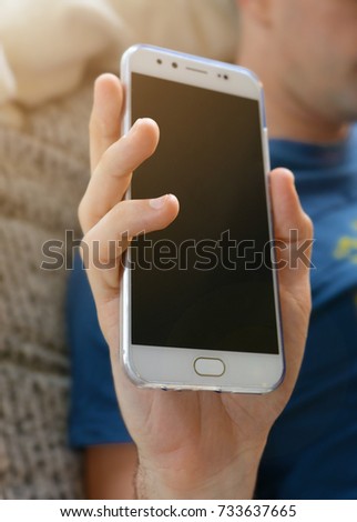 Cropped shot of a mobile phone in a man hand who is relaxing at home sitting on a sofa. Smart phone in a man hand with blurred background and flare light