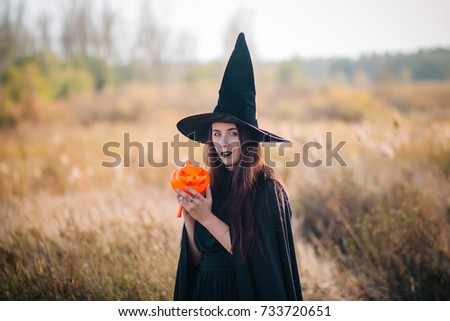 A young witch with pale skin and black lips in a black hat, a dress and a cloak, with a pumpkin in his hands. Girl in a witch costume for Halloween, in the autumn on a background of a field.