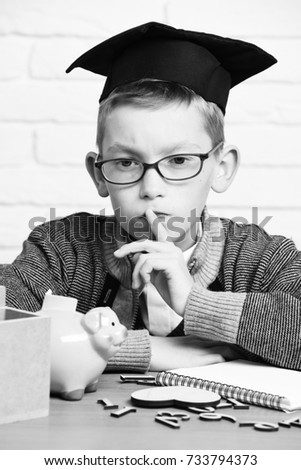 young cute pupil boy in grey sweater and glasses sitting at desk with copybook wooden numbers pink piggy pig bank in graduation cap with raised finger in classroom on white brick wall background
