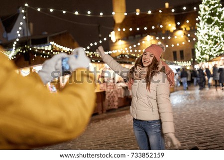 holidays, winter and people concept - happy woman posing for boyfriend with smartphone on square at christmas market