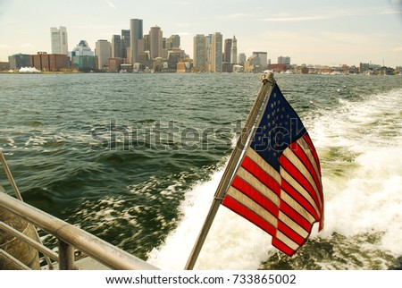 A ferry boat, flagged to the USA, cruises away from the Boston skyline