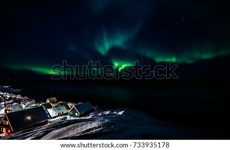 Green glowing of Aurora Borealis with shining stars over the fjord and Inuit village huts, Nuuk, Greenland