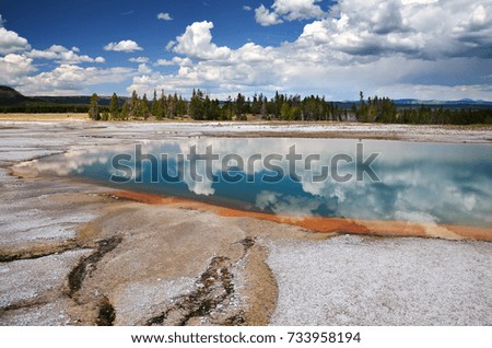 Landscape picture with small rocks and geothermal pool in foreground, reflecting the blue sky and clouds, with some woodland in the background