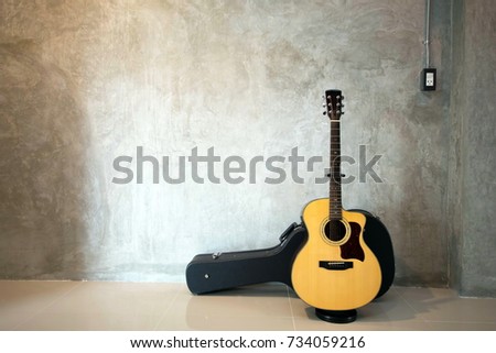 Acoustic guitar on stand with box on the floor and cement wall.