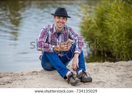 Young smiling man in fashionable clothes sits on the river bank with a phone and listens to music on headphones