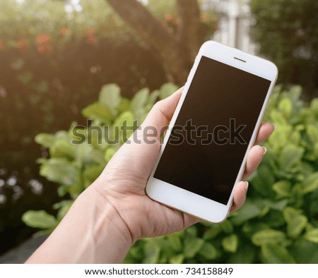 Woman's hand holding a white mobile phone with blank black screen in green garden and lens flare.