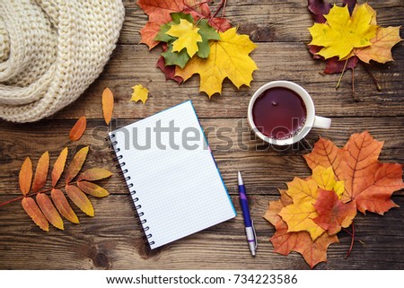 Beautiful autumn picture with yellow, red and orange leaves, a cup of tea, a scarf and a piece of paper with pen on wooden background