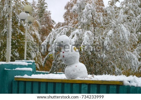Angry, sad snowman for the holiday of Halloween, decorated with red berries of mountain ash. Trees with green and yellow leaves covered with white snow. The first snow in autumn. Winter background.