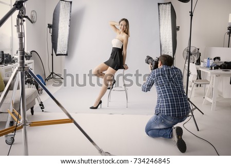 Professional photo shooting at the studio: a beautiful young model is smiling and posing; the photographer is taking pictures with a digital camera