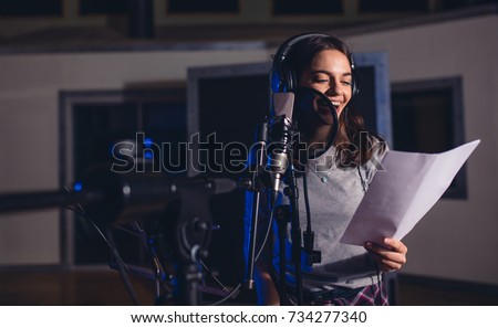 Smiling female singer with microphone and reading lyrics. Woman recording a song in music studio. Female playback singer recording her new album. Royalty-Free Stock Photo #734277340