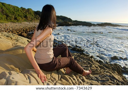 Young girl sits on rocks and looks at sea - sunset light. Shot in Gordon/False Bay, Western Cape, South Africa.