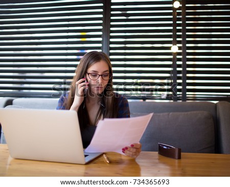 Woman successful magazine editor reading paper documents and taking on mobile phone., sitting in coffee shop. Female skilled writer having cell telephone conversation after work on laptop computer