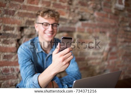 Young smiling hipster guy having video call with friends via mobile phone, sitting against brick wall with copy space. Happy cheerful man manager chatting on cell telephone during work break in office