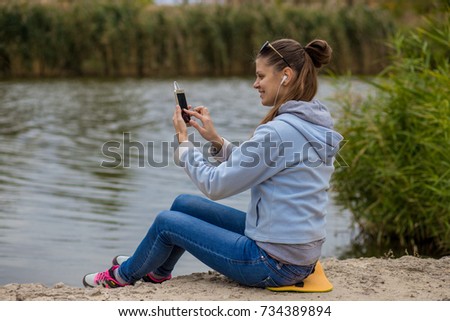 Young woman in excellent spirits sits on the river bank uses a phone and listens to music on headphones