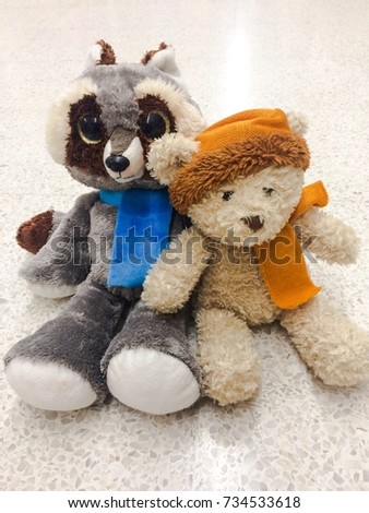  Teddy bear wearing orange yellow color scarf sitting with Raccoon doll wearing blue scarf on white background