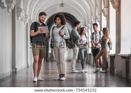 Beautiful young students are resting in university hall, a couple in the foreground is using a digital tablet and smiling Royalty-Free Stock Photo #734541232