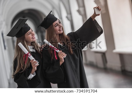 Attractive young female graduates in academic dresses are holding diplomas, doing selfie and smiling while standing in university hall