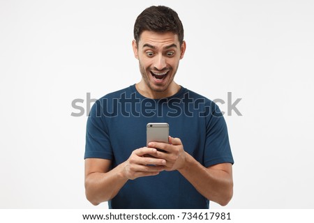 Close up shot portrait of young man isolated on gray background, looking agitated at display of her smartphone smiling and laughing happily, impressed by media content from web