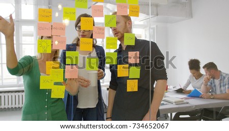 Shot through glass of bright stickers on glass wall and young creative people behind collaborating on new plan ideas. Royalty-Free Stock Photo #734652067