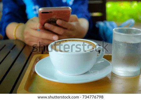 latte coffee in white coffee cup on brown wood table and blur Hand holding Smart phone background