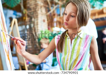Side view portrait of talented teenage girl painting beautiful picture on easel in art class, with other children  in background