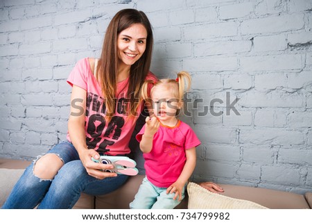 beautiful young mother uses a smartphone to play with a two-year-old daughter sitting on the couch in the room. Dressed in bright colors, colored clothes