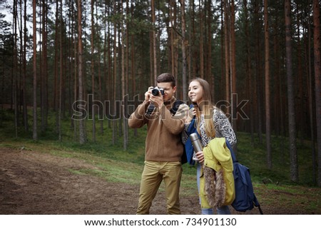 Outdoor portrait of young Caucasian friends boy and girl walking in park or wood together on warm day, male photographer with camera looking for best views and angles, taking pictures of autumn nature
