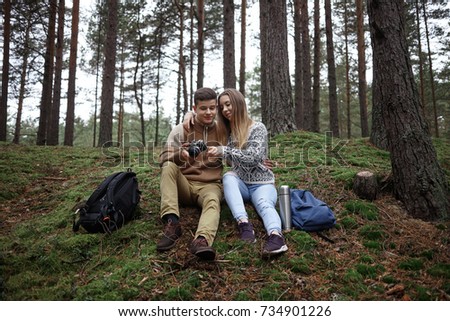 Handsome guy and beautiful girl wearing sensible clothes watching pictures on professional camera, sitting on grass close to each other among trees with backpacks and thermos bottle on the ground