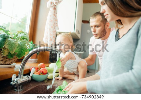 Young beautiful parents and her cute curly toddler daughter washing vegetables together in a kitchen sink getting ready to cook salad for lunch