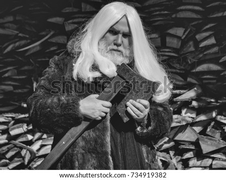 Old man druid with long silver hair and beard in fur coat stands with axe on woodpile background