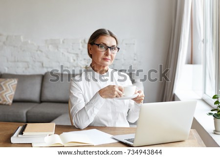 Attractive confident middle aged female psychotherapist in rectangular glasses having coffee during break while working at her desk, engaged in online consulting of adults, children and families