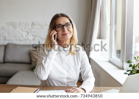 Picture of positive 60 year old female teacher wearing white blouse and stylish rectangular glasses smiling joyfully while talking on smart phone with her grandson during working day at university