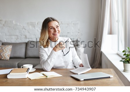 Experienced mature female chief editor holding glasses in one hand and book in other, laughing at sarcastic style of narration while reading novel of popular author. Writing and publishing concept Royalty-Free Stock Photo #734943907