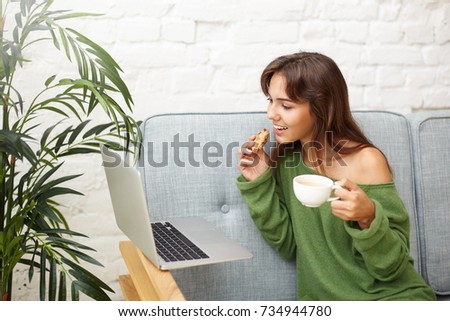 Modern stylish beautiful girl drinking coffee with cookie at home and watching series or movie online using laptop computer, looking at screen with interest. People, technology and leisure concept
