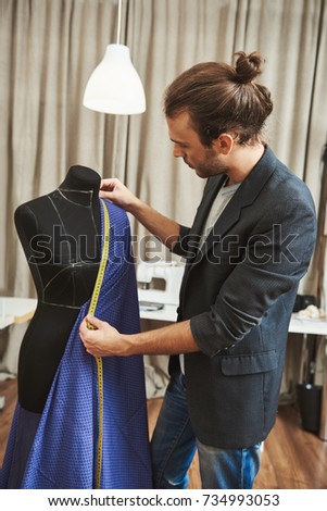 Vertical portrait of adult good-looking caucasian male fashion designer with stylish hairstyle in fashionable outfit in his studio working on new dress for winter clothes collection