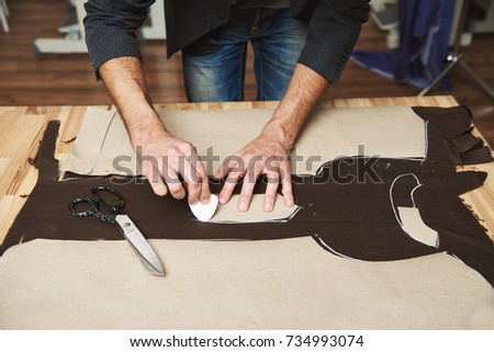 Close cropped view of young professional creative male fashion designer working on new clothes collection, drawing with soap on fabric, cutting out dress parts