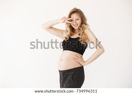 Close up of funny good-looking young europien mom with blond hair in cozy clothes smiling brightfully, posing in studio with white background, holding hand in front of head in V sign, winking, being