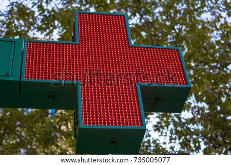 A pharmacy sign, a clinic, a hospital. Sign of the red cross on the street. In the background are the leaves of trees and the sky. Autumn street. The view from the bottom, the background is blurred.