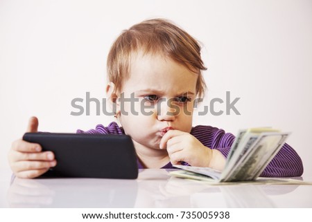 Happy successful business baby girl holding a smartphone in front of bunch of money (humorous picture)